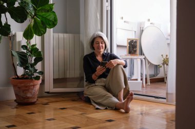 Evde cep telefonu kullanan rahat bir yaşlı kadın balkon kapısının yanında yerde oturuyor.