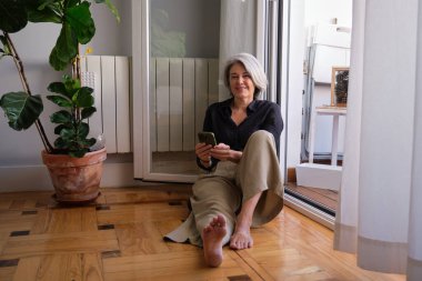 Balkon kapısının yanında rahatlarken cep telefonu kullanan gülümseyen olgun bir kadın, boş zamanların tadını çıkarıyor.