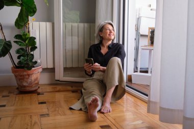 Akıllı telefonu tutan, gülümseyen olgun bir kadın ahşap zemine oturmuş, yukarı bakıyor, güzel bir şeyin hayalini kuruyor.