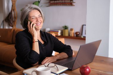Mutlu yaşlı kadın iş için ev telefonu ve bilgisayar kullanıyor.