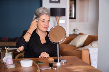 Üst düzey bir kadın yüzüne gua sha taşıyla masaj yapıyor, evdeki ahşap masadaki aynaya bakıyor.