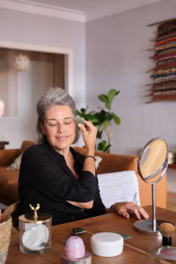 Olgun bir kadın yüzüne gua sha yeşim taşıyla masaj yapıyor, evde kendine bakmanın keyfini çıkarıyor.