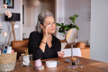 Makyaj malzemelerini çıkarmak için pamuk ped kullanan olgun bir kadın. Elinde ayna ve güzellik ürünleri olan ahşap bir masada oturuyor.