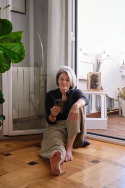 Evde tahta zeminde çıplak ayakla otururken cep telefonu kullanan rahat olgun bir kadın.