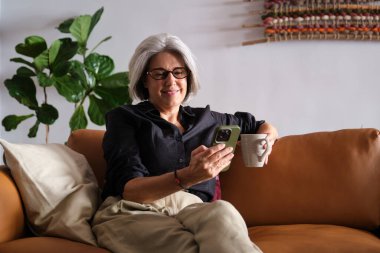 Mutlu olgun bir kadın rahat koltukta rahat rahat oturup cep telefonu kullanıyor ve kahve içiyor.