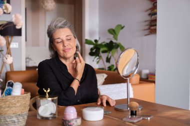 Olgun bir kadın yeşim taşıyla evde yüzüne masaj yapıyor, cildine güzellik tedavisi uyguluyor.