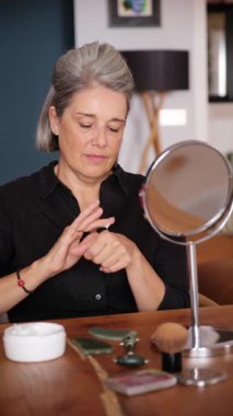 Kıdemli kadın yüzüne nemlendirici sürüyor, aynaya bakıyor.