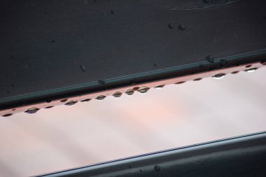 Karanlık bir yüzeyden sarkan su damlacıklarının makro görüntüsü ve altında ince pembe bir yansıma var.