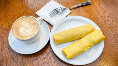 Ahşap bir kafe masasında bir fincan sıcak kapuçinoyla servis edilen nefis rulo krepler.