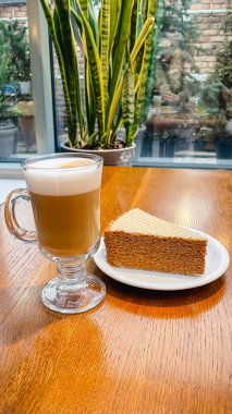 Cam bardakta katmanlı latte ve bir dilim waffle keki, üzerinde konsantre süt olan ahşap bir kafe masasında.