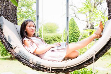 Modern bir dizüstü bilgisayarla hamakta yatarken kameraya gülümseyen ve uzaktan evde çalışan, serbest çalışan mutlu bir genç kadın..