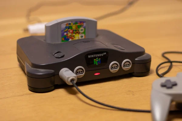 Bir Nintendo 64 konsolu. Şarjör yuvasında ışık açık ve Super Mario 64 ve fişe takılı bir Controller var.
