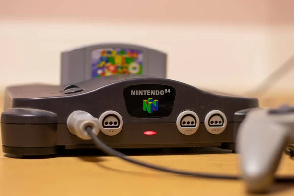 Bir Nintendo 64 konsolu. Şarjör yuvasında ışık açık ve Super Mario 64 ve fişe takılı bir Controller var.