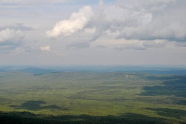 Dağ bölgesinin panoramik manzarası. Dağların üzerinden geçen beyaz bulutlar, yeşil ormandaki gölgelerini yansıtıyor..