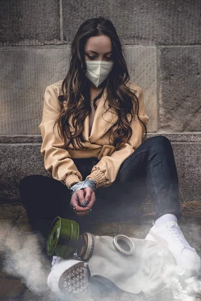 女性身に着けています保護顔マスクでザ コヴィト 19パンデミック中に手で縛られましたテープで地面に座っています古いヴィンテージ戦時ガスマスク毒性の煙の雲の中に — ストック写真