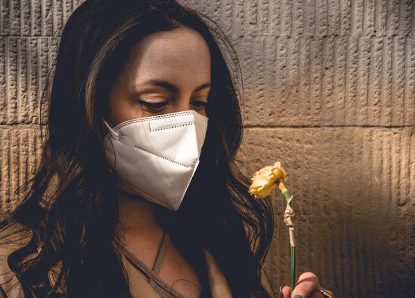 Covid 19パンデミックの間にフェイスマスクを身に着けている若い女性のムーディーの肖像影の壁に対して彼女の手に枯れて春の花を保持 — ストック写真