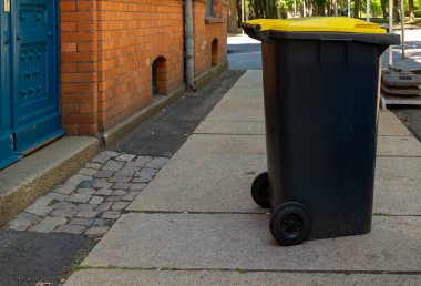 A black trash bin with a bright yellow lid sits on a smooth sidewalk beside a red brick building. Trees provide shade in the background, indicating a calm, sunny day.
