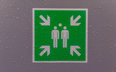Yeşil tabela yağmurlu bir ortamda acil çıkışları işaret ediyor. Damlacıklar, aciliyet ve güvenliği vurgulayarak olay yerine pırıl pırıl bir etki katıyor..