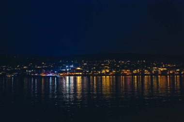 Karanlık gökyüzünün altında, liman manzarası suyun yüzeyinde dans eden canlı ışıklarla güzel bir şehir manzarası sergiliyor. Huzurlu bir sahne sükunet ve içgözlem davet eder..