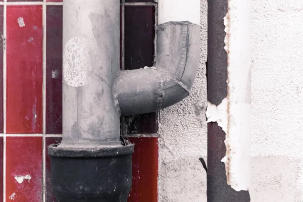 Gri bir boru eğilir ve bir duvardaki siyah boruya bağlanır. Duvarda yıpranma ve yıpranmayı gösteren kırmızı ve beyaz fayanslar var. Şehir ortamında açık bir gün..