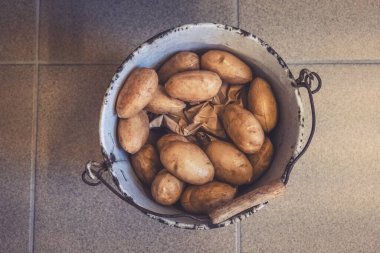 Sıcak bir mutfakta, metal bir kova yeni toplanmış patateslerle dolu fayanslı bir zeminde oturur. Yumuşak ışık toprak tonlarını aydınlatır ve rahat bir atmosfer yaratır..
