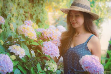 Bahçesinde pembe ortanca çiçekleri olan hasır şapka giyen Asyalı kadın.