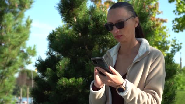 一个女人在大自然的背景下翻阅着智能手机的显示屏 一个戴墨镜和手持手机的女人的特写镜头 手指触摸着手机的屏幕 — 图库视频影像
