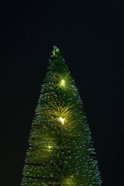 Siyah arkaplandaki yapay ağaç. Ağaçta şenlikli sarı fenerler yanıyor. Fotoğraf aşağıdan yukarıya doğru çekilmiş. Ağacın kendisi de çok büyük görünüyor..