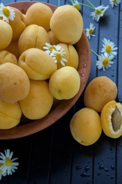 Olgun kayısılarla dolu eski bir kil tabağı siyah ahşap bir arka planda duruyor. Yakın planda çekilmiş. Kayısılar küçük papatya çiçekleriyle süslenir..