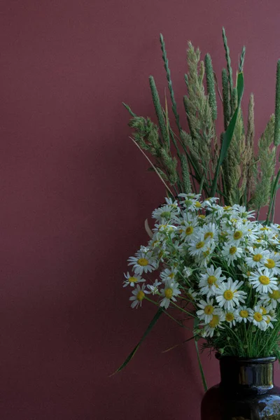 a bouquet of wild flowers in a vase of dark glass stands on a burgundy background. close-up. photo is suitable for a menu or a postcard. there is free space for text