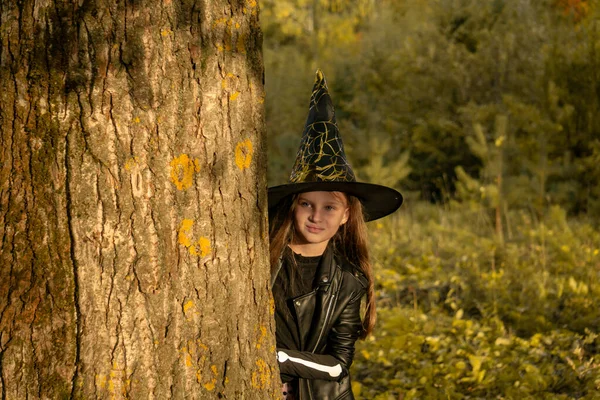 Vicces kislány boszorkányjelmezben Halloweenkor egy nagy fa mögül néz ki. Boldog Halloweent!. — Stock Fotó