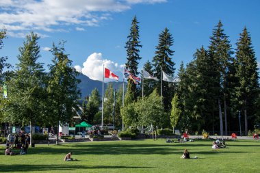 Whistler, Kanada - 5 Temmuz 2020: Whistler Village 'da bayrak sallayan direklerin görüntüsü. İnsanlar güneşli bir günde yeşil çimlerde oturuyorlar.