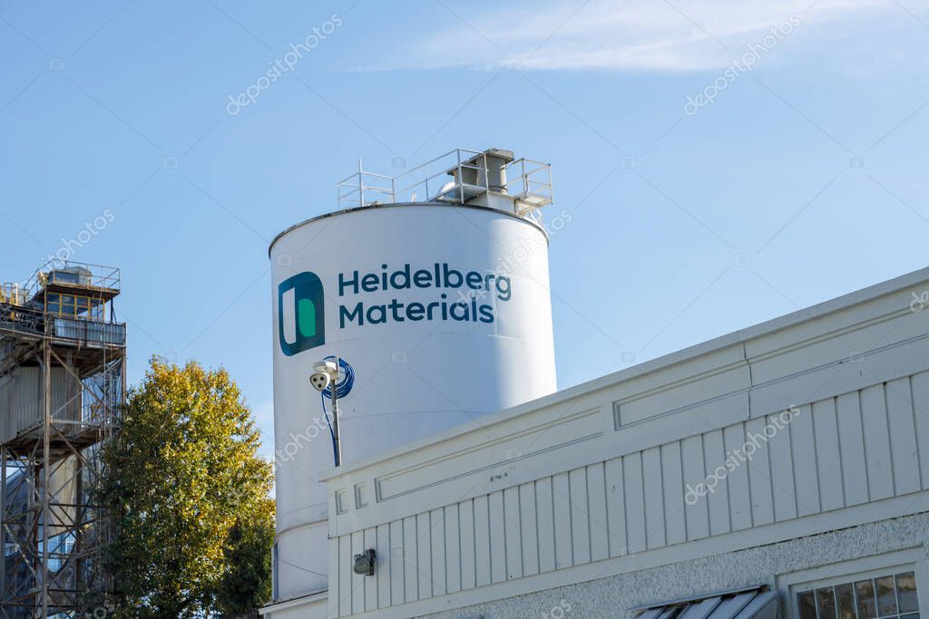 Vancouver, Canada - Oct 6,2025: Heidelberg Materials white cylindrical storage silo with company logo and platform on top beside industrial building and processing tower