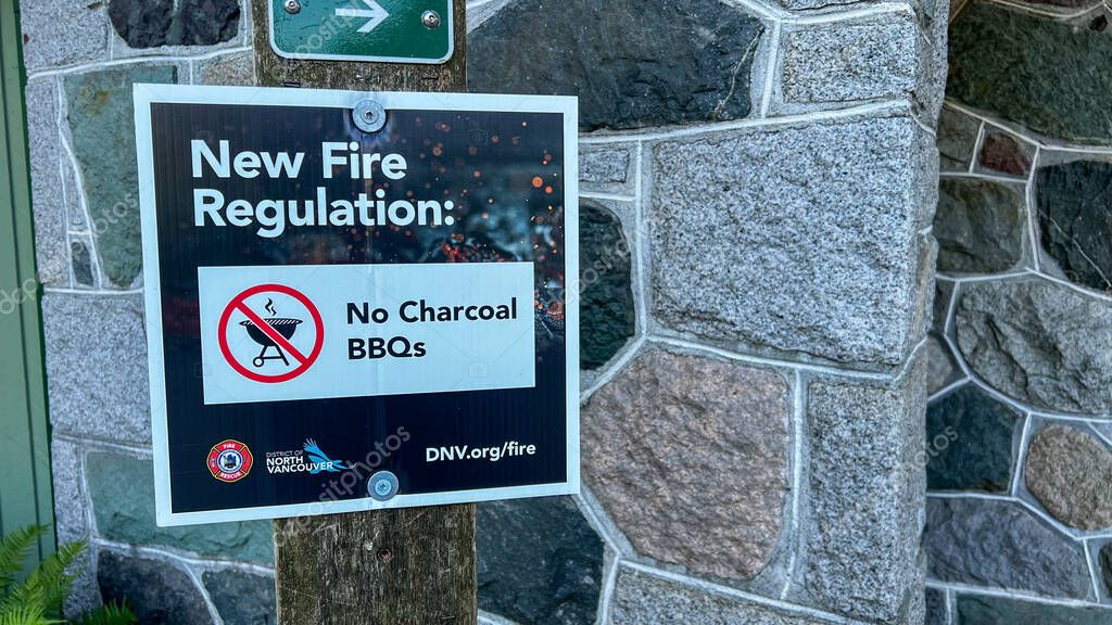 Vancouver, Canada - Sep 19,2025: District of North Vancouver fire regulation sign prohibiting charcoal BBQs mounted on wooden post against stone wall