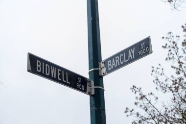 Vancouver kavşağındaki yeşil sokak işaretleri Bidwell Caddesi 900 blok ve Barclay Caddesi 1600 blok bulutlu gökyüzü ve ağaçlarla kaplı karanlık direğe monte edilmiş.