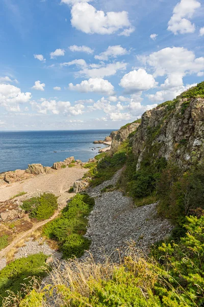 Danimarka 'daki Hovs Hallar Doğa Rezervi' nde dramatik bir kıyı manzarası: engebeli uçurumlar, kayalık plaj ve mavi gökyüzü altında beyaz bulutlar, tepe boyunca yeşillik.