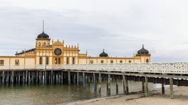 Varbergs kallbadhus, büyük yuvarlak bir gül pencereli, çarpıcı sarı bir hamam olan ahşap bir iskelenin sonunda duruyor. Beyaz korkuluklar geçidi çerçeveletiyor, mimari, seyahat ve sahil cazibesini aktarıyor.