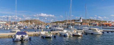 İsveç 'in Tjorn Adası' ndaki güneş ışığı limanında yatlar, yelkenliler, sahil boyundaki pastel evler ve tepede parlak mavi bir gökyüzü bulunan sakin bir marina bulunur..