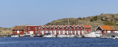 Tjorn Adası 'ndaki Ronnang' da sakin bir İsveç limanında renkli kırmızı ahşap Stuga kulübeleri, ahşap bir rıhtım ve berrak mavi bir gökyüzünün altında usulca sallanan yelkenliler bulunur..