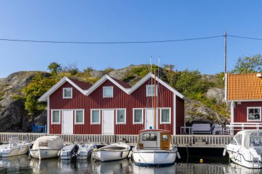 Tjorn Adası 'ndaki Ronnang' da sakin bir İsveç limanında renkli kırmızı ahşap Stuga kulübeleri, ahşap bir rıhtım ve berrak mavi bir gökyüzünün altında usulca sallanan yelkenliler bulunur..