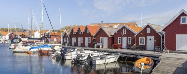 Tjorn Adası 'ndaki Ronnang' da sakin bir İsveç limanında renkli kırmızı ahşap Stuga kulübeleri, ahşap bir rıhtım ve berrak mavi bir gökyüzünün altında usulca sallanan yelkenliler bulunur..