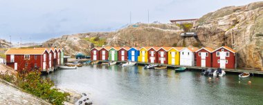 İsveç 'in Vastra Gotalands kentindeki Smogen limanında engebeli kayalara karşı kurulu renkli ahşap kayıkhanelerin panoramik manzarası. Küçük tekneler parlak bir gökyüzünün altında hareketsiz bir kıyı sahnesi oluşturarak durgun sularda sürüklenirler..