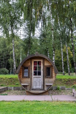 Uzun huş ağaçları tarafından çerçevelenmiş, huzurlu bir orman parkında, rahat, yuvarlak, ahşap bir tatil kulübesi bulunuyor. Kırsal kapılar ve küçük pencereler, kırmızı perdelerle, doğada büyüleyici, samimi bir geri çekilme yaratır..