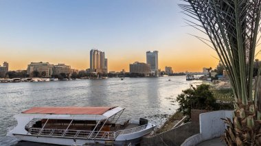 Günbatımında Kahire 'de sakin bir Nil nehri manzarası, modern bir ufuk çizgisi, kıyıya demirlemiş gölgeli bir nehir teknesi, ön planda palmiye ağacı ve suya yansıyan ılık turuncu ışık..