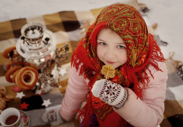 A girl in a Russian headscarf sits on the bank of the river with a samovar, bagels and drinks tea