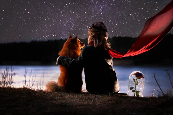 Kırmızı eşarplı siyah paltolu bir çocuk nehir kıyısında kırmızı bir köpekle oturur ve gökyüzüne bakar.