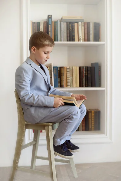 Takım elbiseli çocuk sandalyede otururken kitap okuyor.