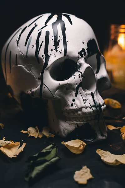 Decorative white human skull in a mysterious setting with burning candles and dry flower petals, black background, close-up, vertical orientation