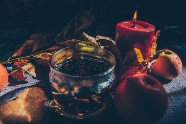 Bitki çaylı bir cam kadeh olgun bir elma ve yanan bir mum yanında duruyor. Arka planda kuru sarı sonbahar yaprakları var.