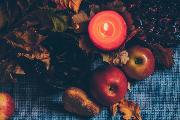Autumn picture with a lit candle, pastries in the form of a heart, ripe sweet apples and dry leaves, close-up, top view